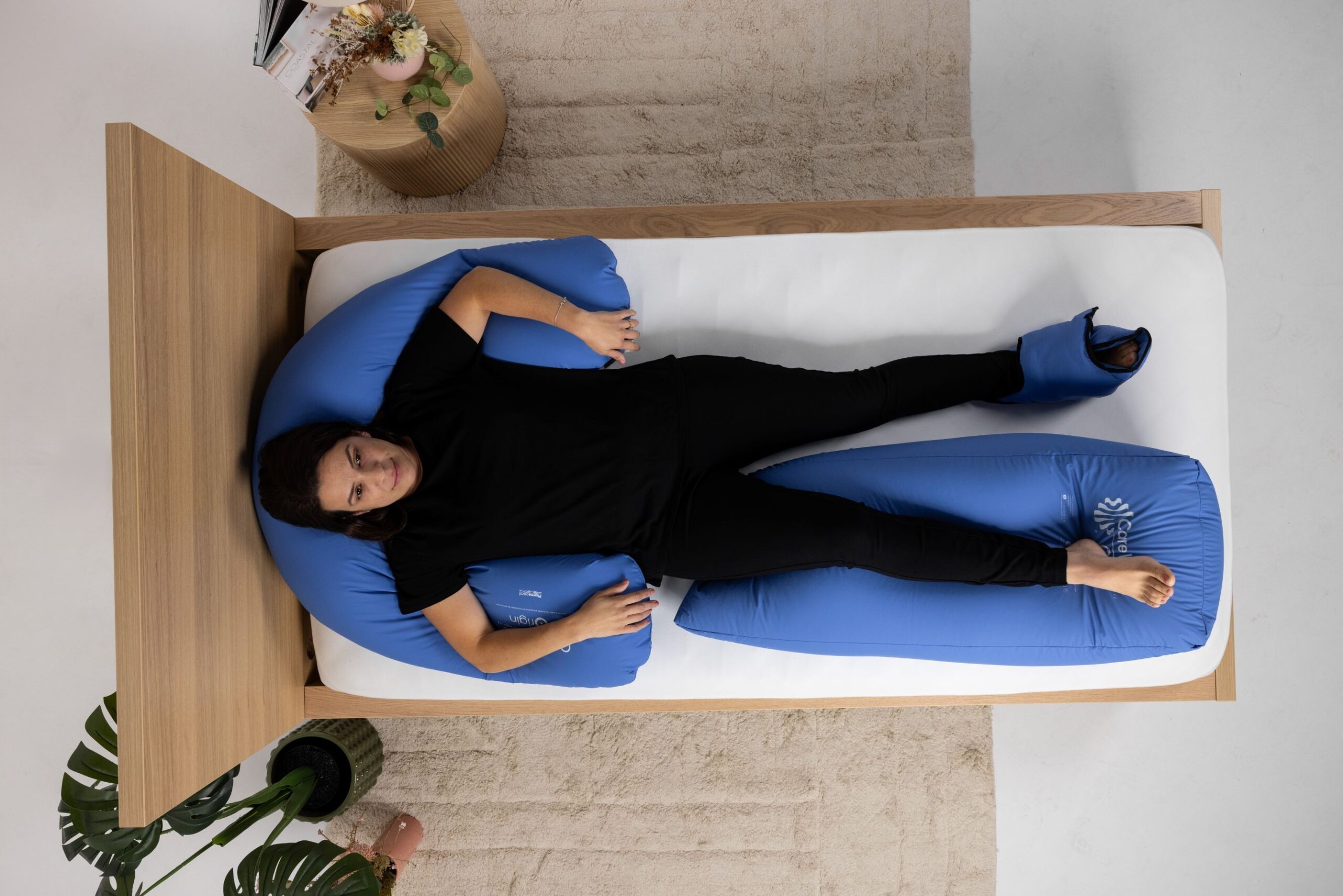 Aerial of a CareWave blue half-moon pillow, CareWave blue XXL abduction cushion and CareWave blue bootie with user on a wooden bed. Brown rug, indoor plants and bedside table in background.