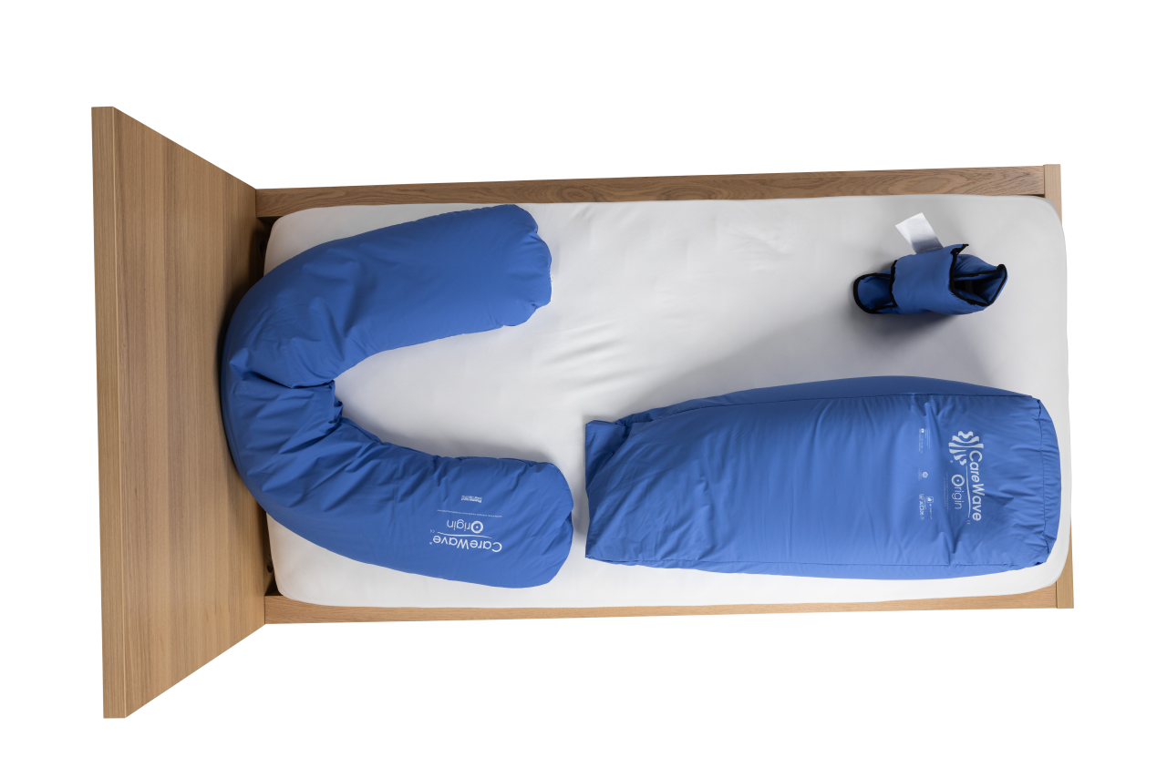 Aerial view of wooden bed, with bedhead to the left. A CareWave blue half-moon pillow, CareWave blue XXL abduction cushion and CareWave blue bootie sit on the bed. Aerial of a CareWave blue half-moon pillow, CareWave blue XXL abduction cushion and CareWave blue bootie on a wooden bed.