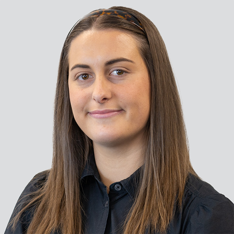 Headshot of young woman named Grace Mascull, smiling into camera. Brown hair, brown eyes, black blouse, grey background.