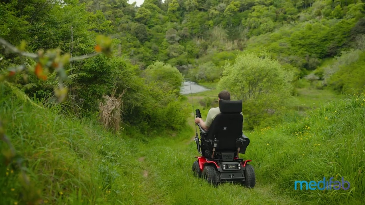 Rear view of man named Phillip driving his Magic Mobility XT4 Four Wheel Drive Electric Wheelchair down a grassy trail, surrounded by trees. A blue Medifab logo sits in the bottom right corner of the image.