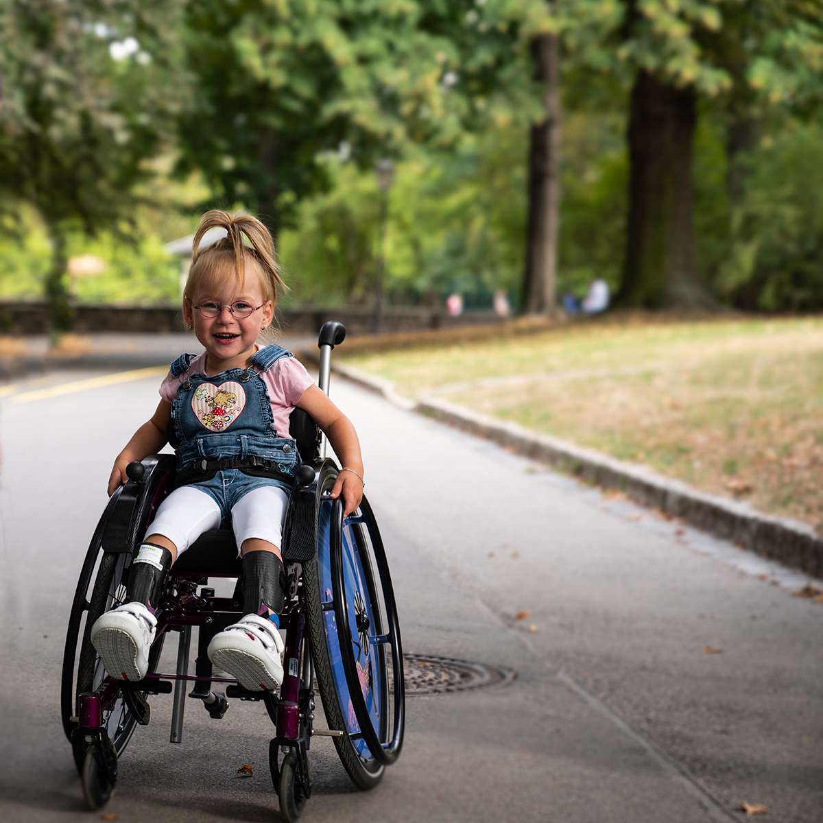 Small girl in a manual wheelchair with large wheel rims and smiling at the camera on a path.