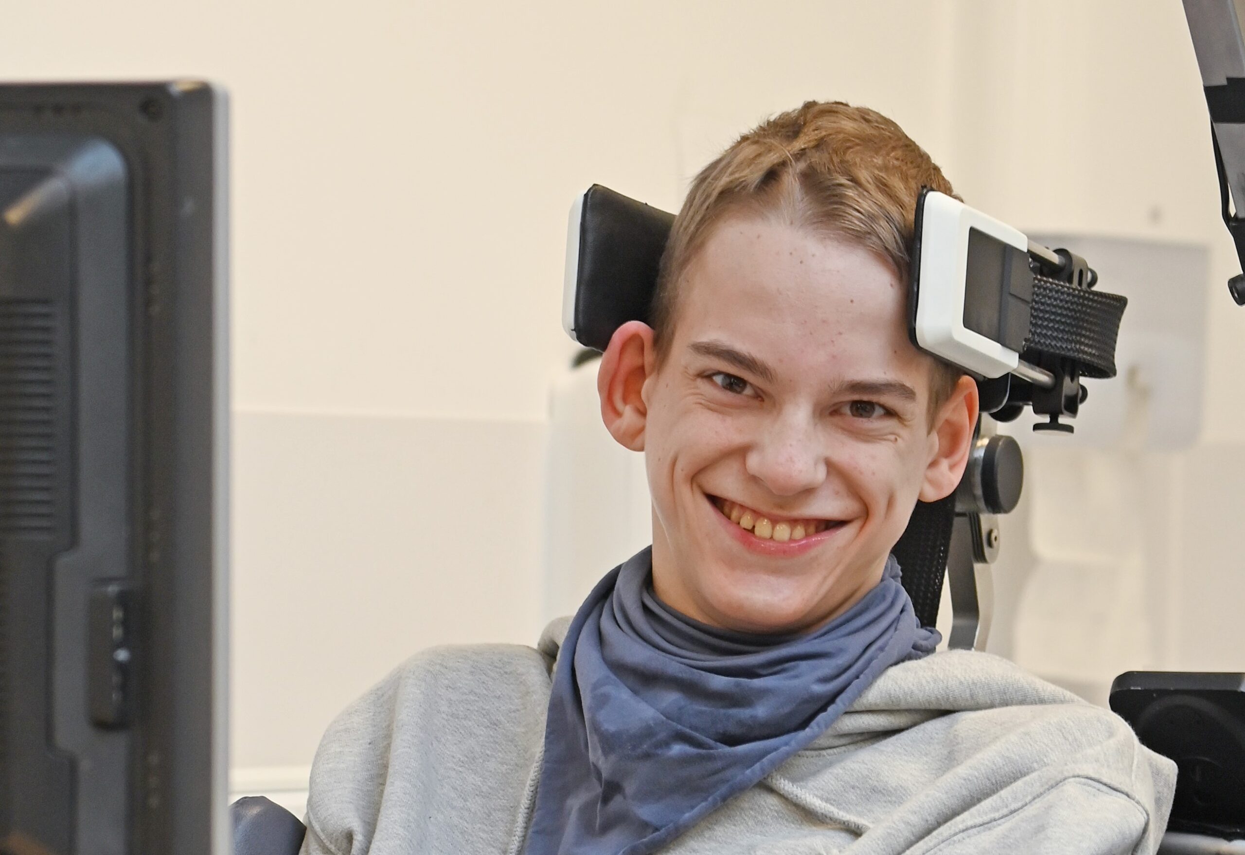 A young boy sits in a wheelchair and smiles at the camera. The shot is from the chest upwards, and he has the CoMoveIT Wheelchair steering system attached to his head to help him steer.