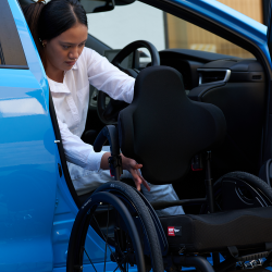 Young women, Chelsea, sitting in the drivers seat of a blue car about to get into her wheelchair.