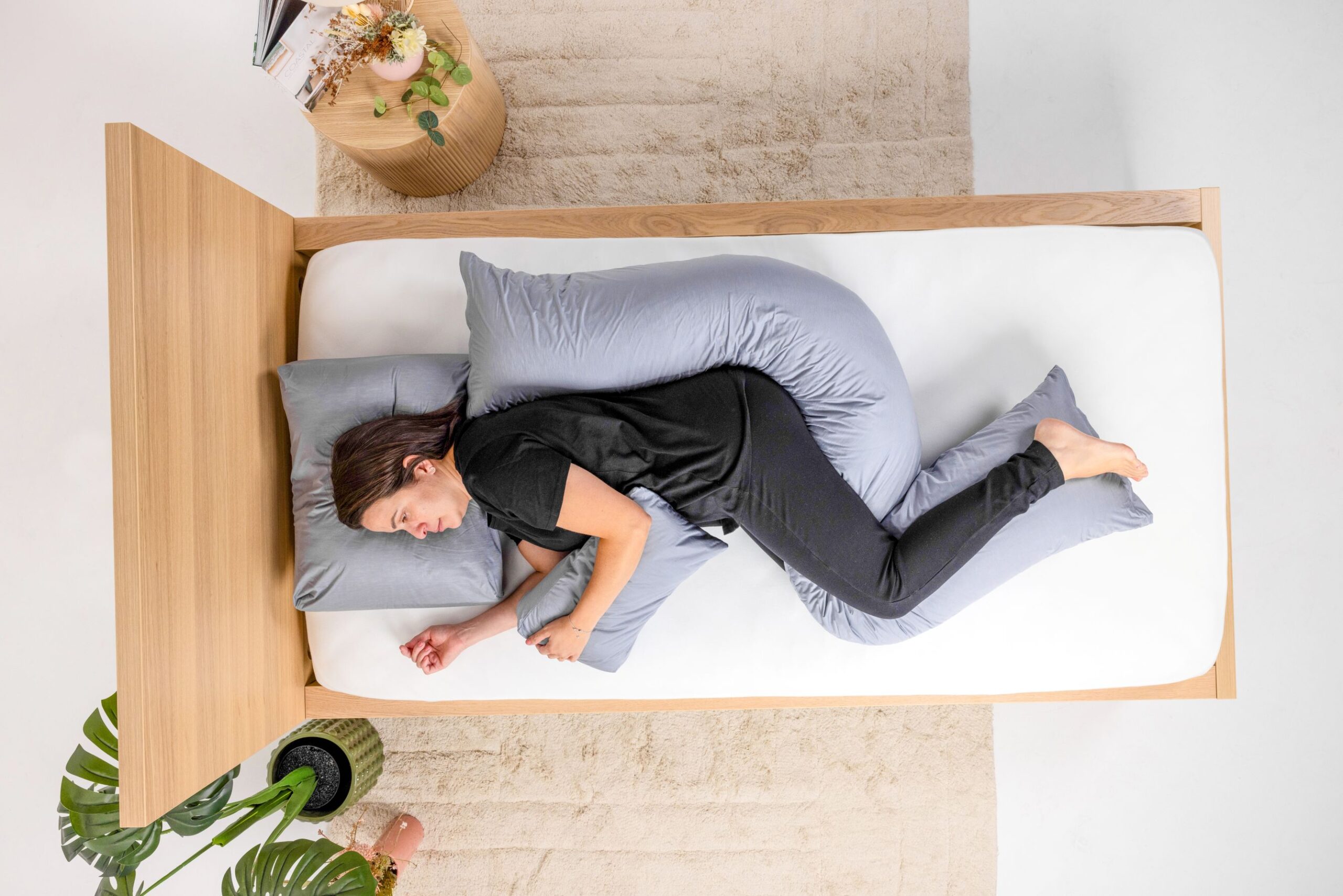 Aerial view of a woman dressed in black lying on a wooden bed with a white mattress and grey occipital, decubitus and hand wedge postural support cushions. Brown rug, indoor plants and bedside table in background.