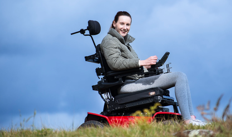 Side on image of a young woman in an power wheelchair on some grass, smiling against a blue sky
