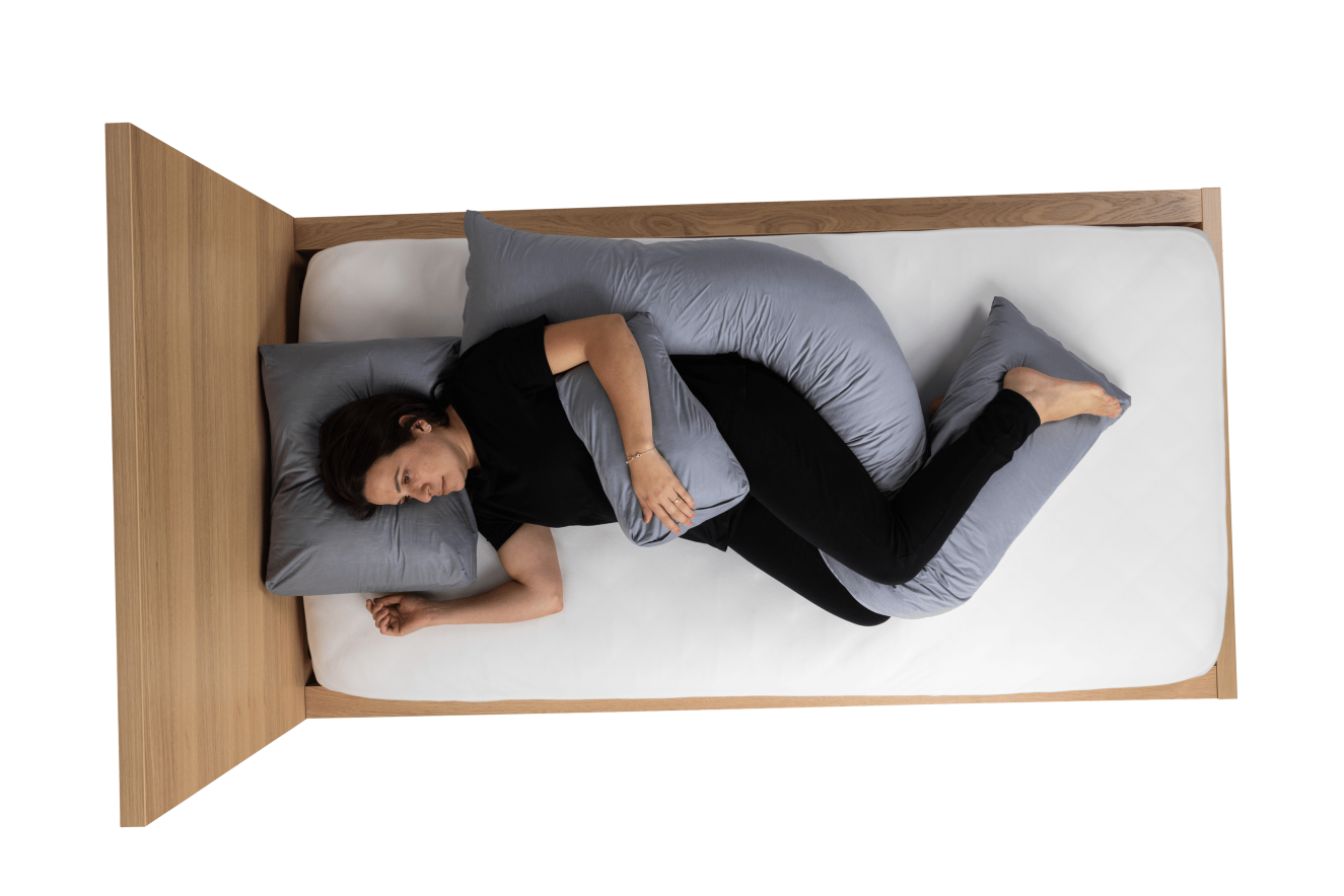 Aerial view of a woman in black clothing lying on a wooden bed with a white mattress, supported by grey Poz'In'Form postural support cushions - specifically an occipital cushion under her head, a hand wedge cushion supporting her left hand on her stomach, and a decubitus cushion supporting her back and legs.