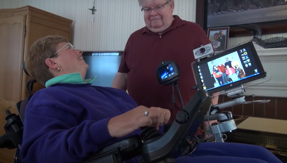 Gae Skager in a powered wheelchair using a Mount’n Mover arm to hold a tablet and secondary device, allowing for accessible communication and computing. The mount is securely attached to her wheelchair and positioned for easy viewing and interaction, while a man stands nearby engaging with her.