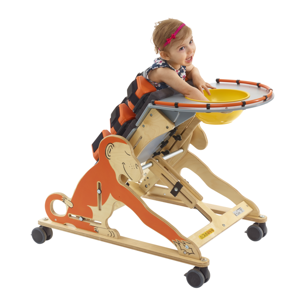 A grinning little girl standing with assiatance from a Jenx monkey prone stander,  playing with a bowl in the attached tray table.
