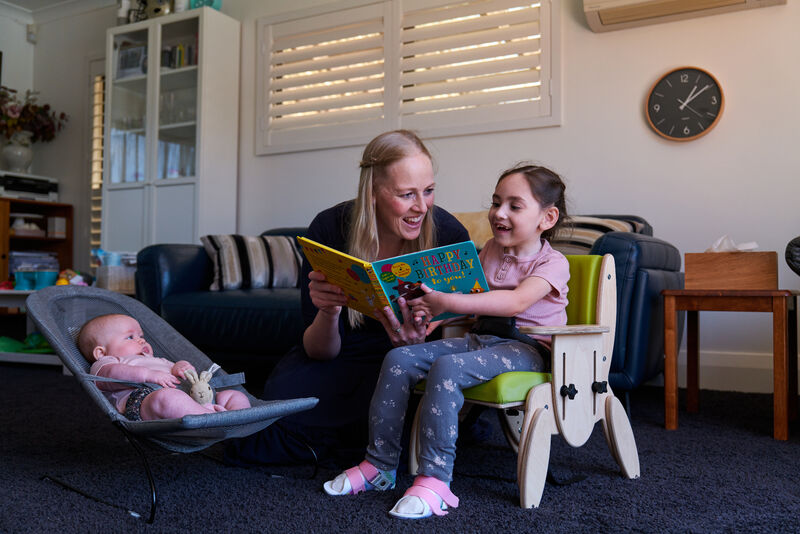 A young girl sitting in a small indoor green chair, with a baby in a rocker beside her, whilst her mum reads her a picture book in a living room.