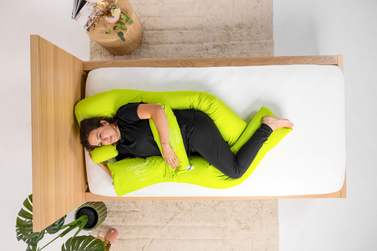 Aerial of a CareWave green body bolster postural cushion, CareWave green decubitus postural cushion and a CareWave green cylindrical postural cushion with user on a wooden bed. Brown rug, indoor plants and bedside table in background.